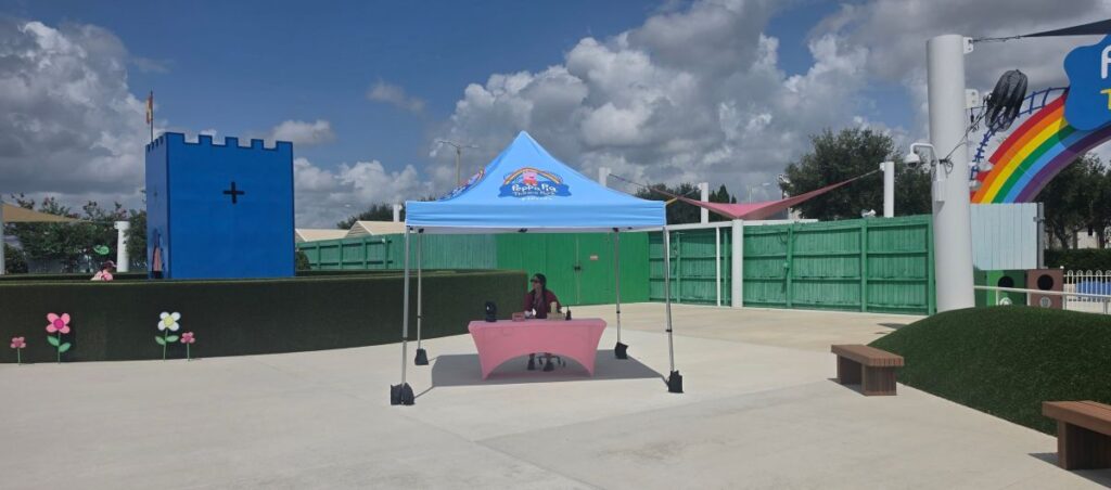 Blue pop-up tent over a pink table at an outdoor playground; a person sits behind it with a rainbow arch in the background.