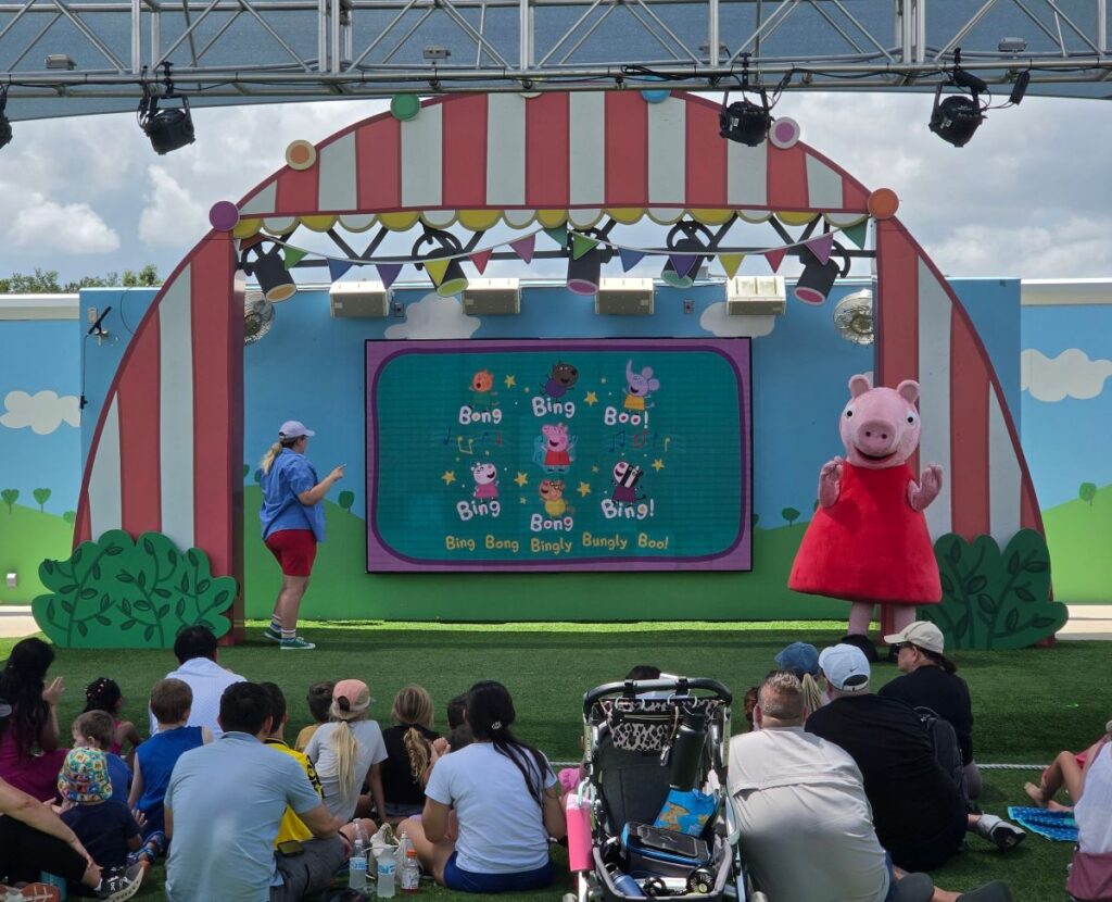 Outdoor kids' stage with a colorful screen showing cartoon characters and the words 'Bing Bong'; a pink pig mascot in a red dress stands to the right while children sit on the grass in front.