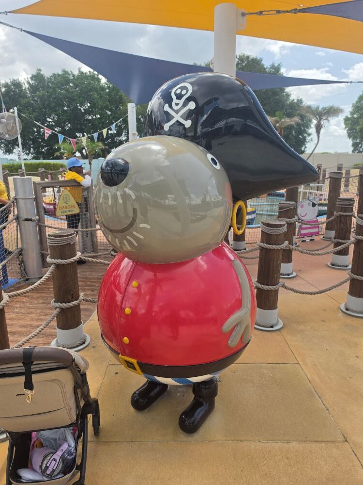 Giant cartoon pirate pig statue, wearing a red vest and black boots, with a black pirate hat and skull-and-crossbones, at a sunny outdoor play area.