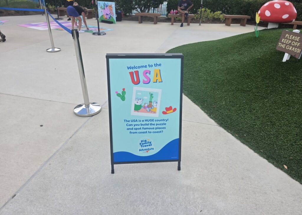 Outdoor freestanding sign at Peppa Pig Theme Park advertising a USA-themed activity, with 'Welcome to the USA' and a cartoon image on a blue panel.
