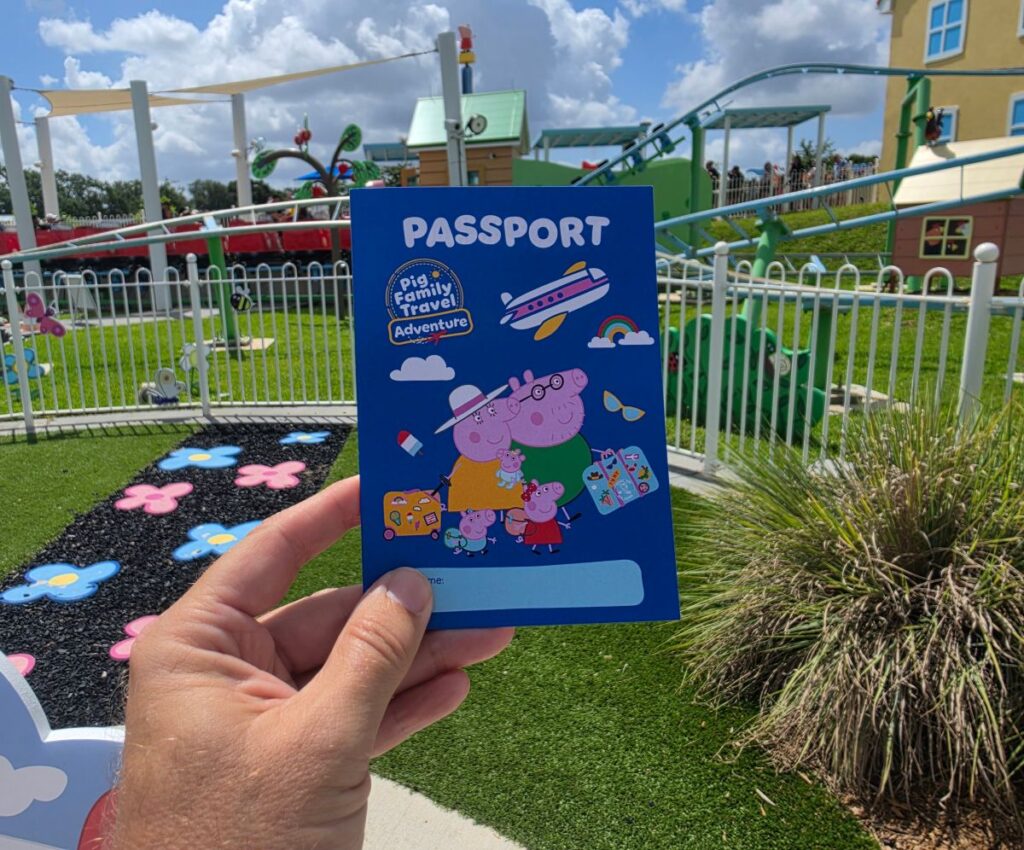 Blue passport card featuring Peppa Pig family travel artwork held up in a colorful playground setting at Peppa Pig Theme Park