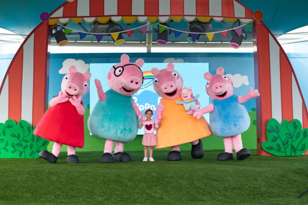 Child posing with four Peppa Pig characters on a colorful stage backdrop at a kids' event. promotional photo for Pig Family Travel Adventure