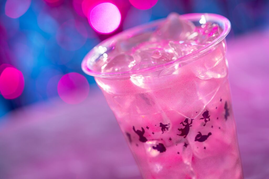 Close-up of a pink iced drink in a clear plastic cup with ice, set against blue and magenta bokeh lights.