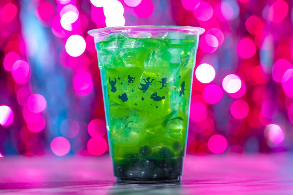 Clear plastic cup with a bright green, ice-filled drink; pink bokeh lights fill the background, cup decorated with black fairy silhouettes.