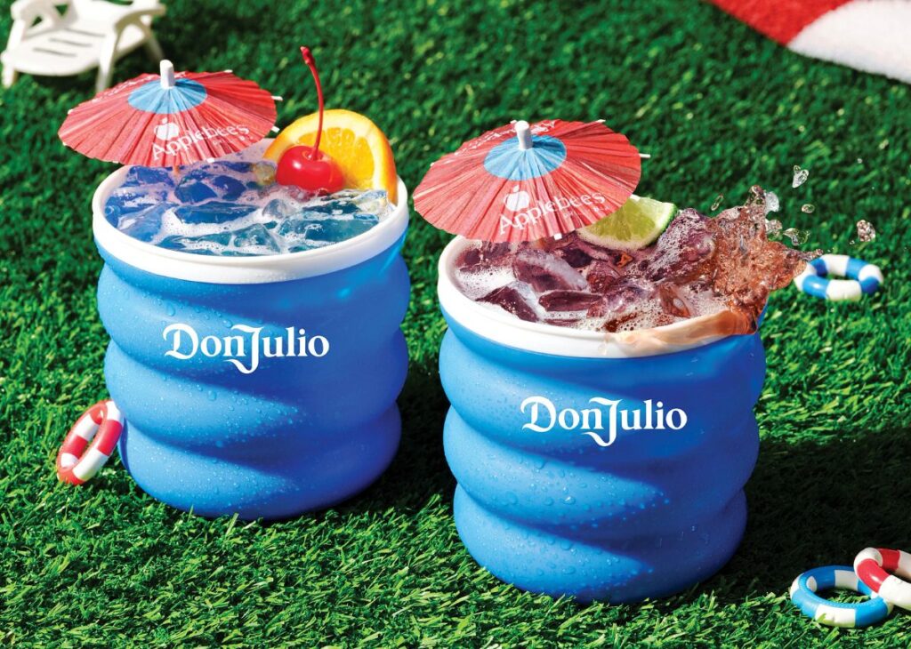 Two blue Don Julio drink tumblers on green grass, filled with ice and garnished with fruit and red umbrellas on top.