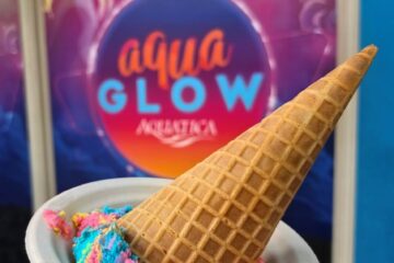 Handheld waffle cone with pink, blue, and yellow scoops in a white cup, in front of a colorful Aqua Glow sign on a blue wall for an AquaGlow food review