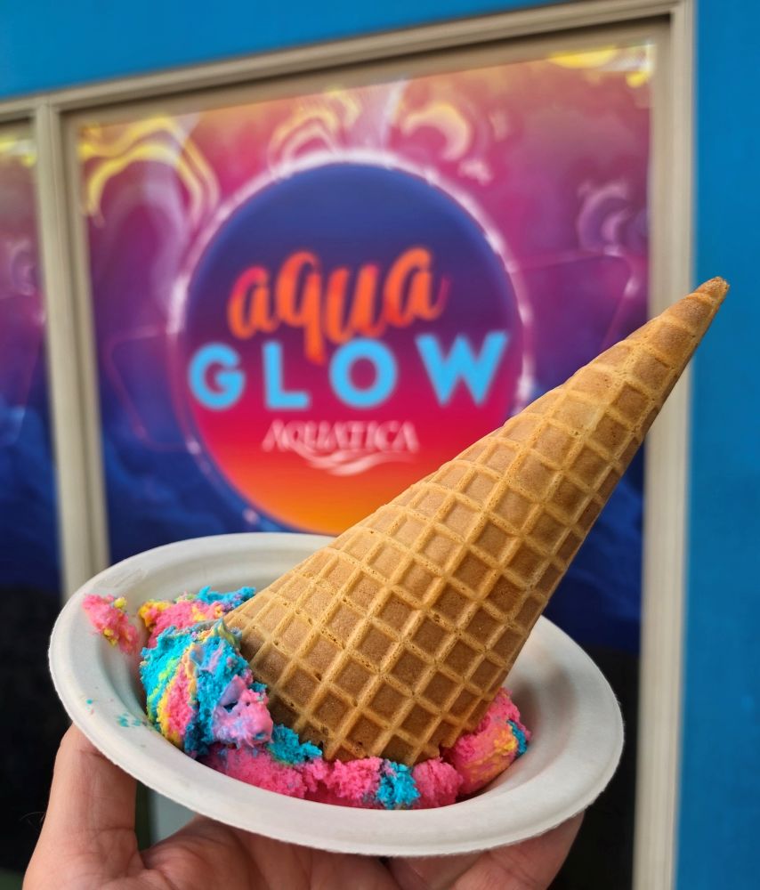 Handheld waffle cone with pink, blue, and yellow scoops in a white cup, in front of a colorful Aqua Glow sign on a blue wall for an AquaGlow food review
