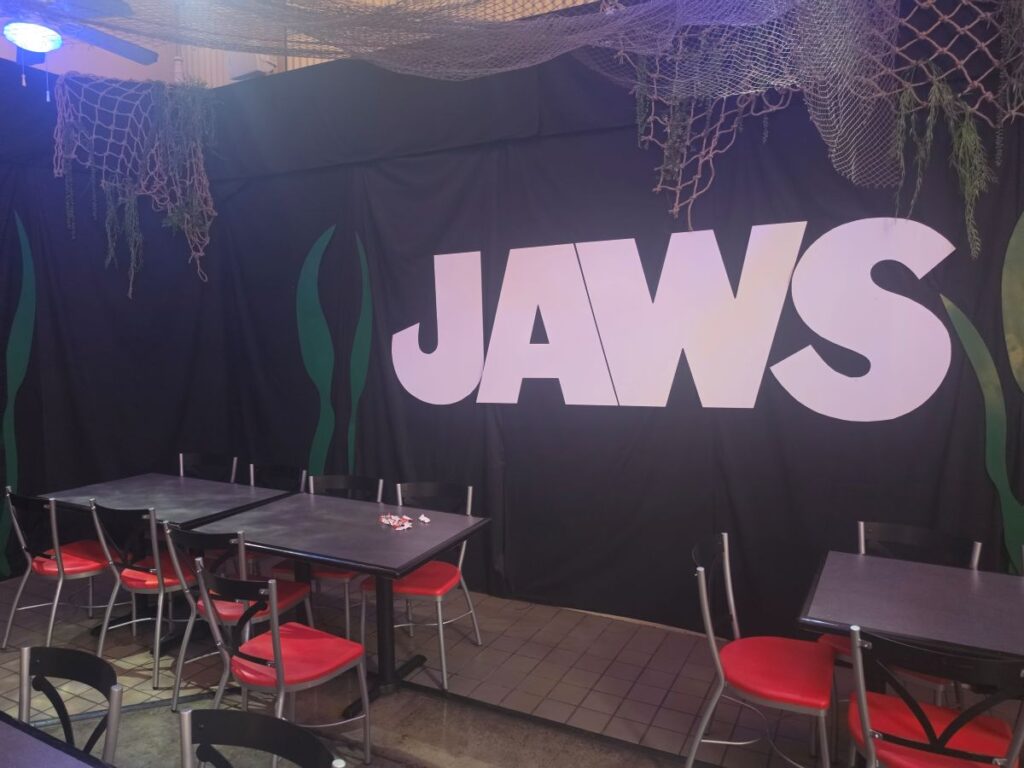Cafe-style event space with a large white 'JAWS' logo on a black backdrop, green seaweed shapes, and nets hanging from the ceiling; metal tables and red-cushioned chairs are arranged in rows.