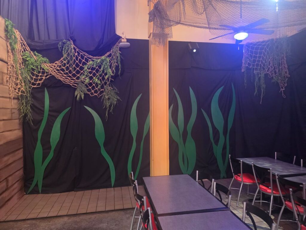 Dining area with black backdrop featuring large green seaweed shapes, hanging nets, and a wooden wall on the left; tables and red-cushioned chairs nearby.