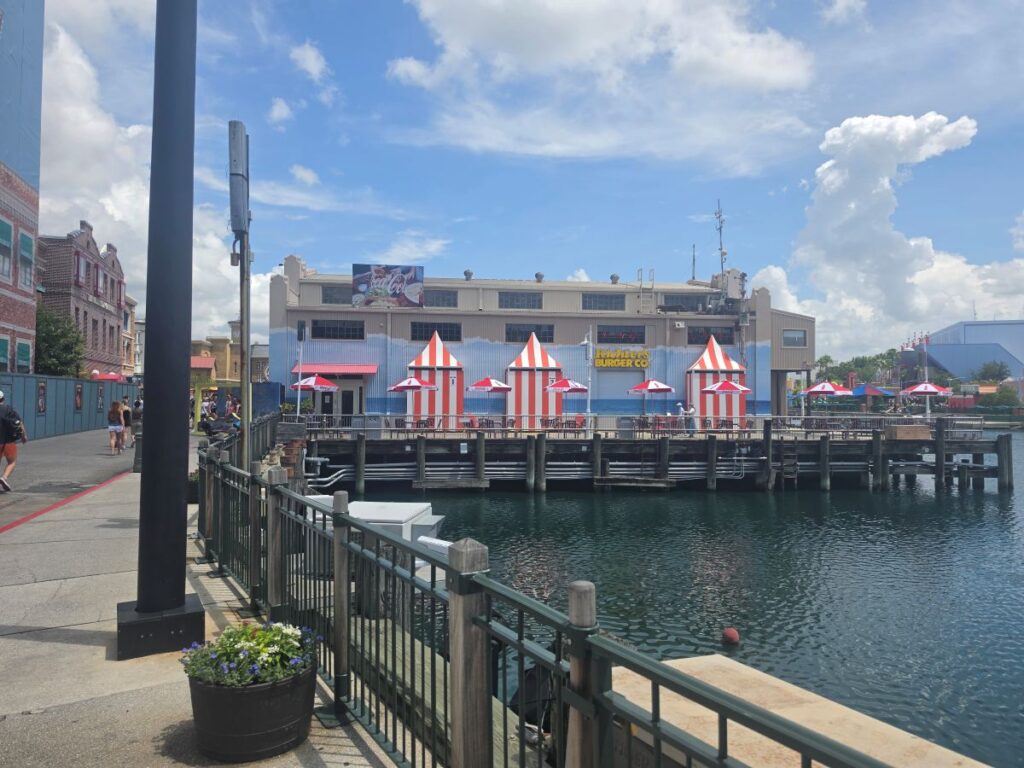 Boardwalk with red-and-white striped tents along a blue waterfront building and seating, overlooking calm water.