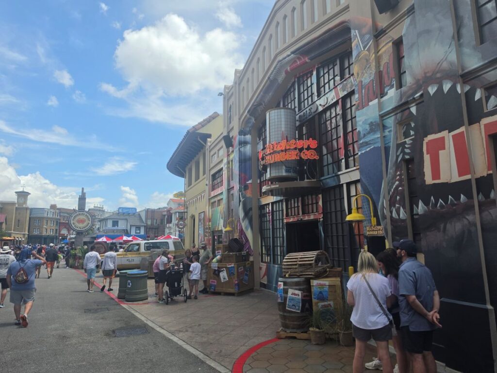 Crowded outdoor shopping street with colorful mural buildings, a 'Richter's Burger Co.' sign, and people browsing food stalls.