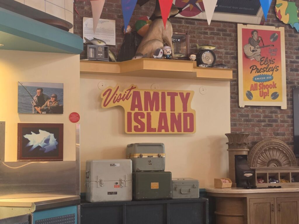 Display area with a bold 'Visit Amity Island' sign, vintage suitcases, and retro posters on a brick-wall backdrop.