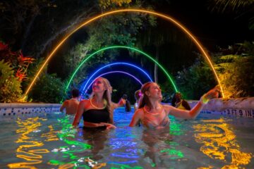 Group of people dancing and smiling in a pool at night under glowing neon rainbow arches over the water at AquaGlow Aquatica Orlando