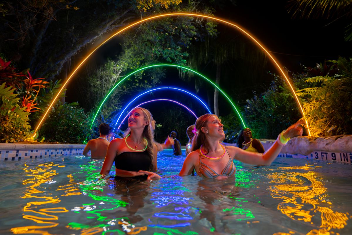 Group of people dancing and smiling in a pool at night under glowing neon rainbow arches over the water at AquaGlow Aquatica Orlando