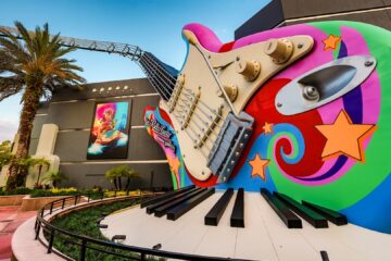 Gigantic colorful guitar sculpture with a piano keyboard at the base, set beside palm trees and a modern building entrance.