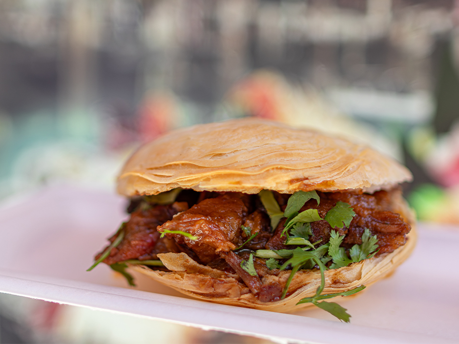 Braised meat sandwich with cilantro in a flaky bun on a white platter, close-up.