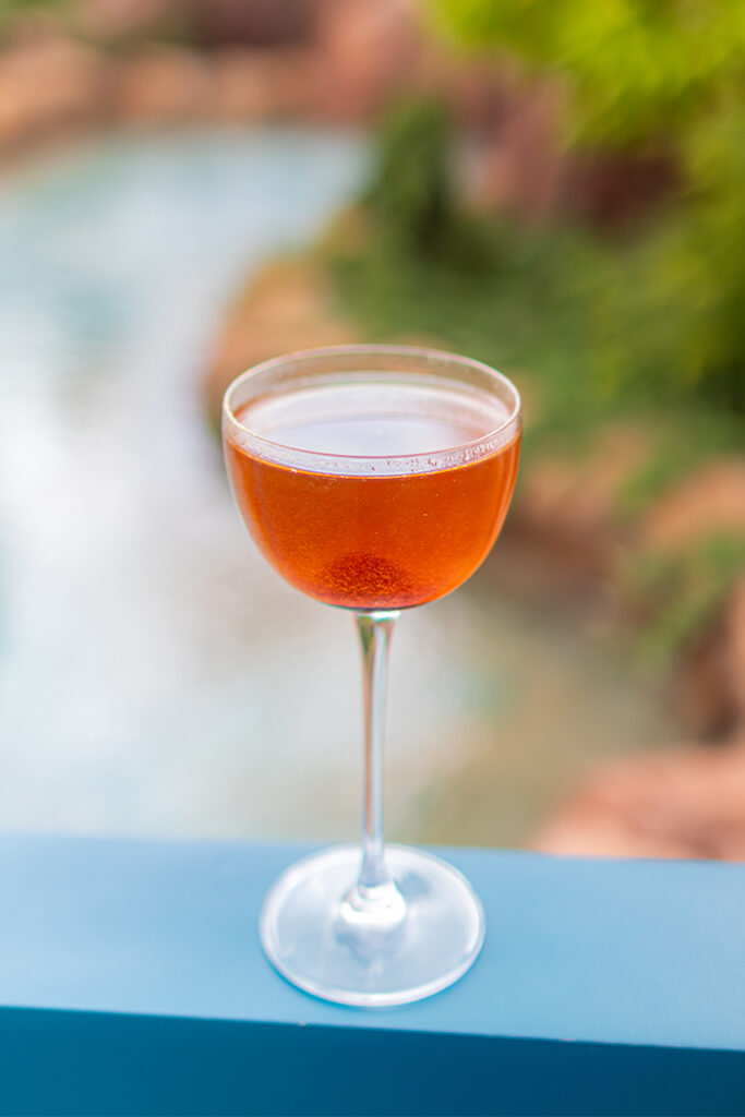 A stemmed glass with an orange beverage on a blue railing, blurred garden backdrop behind it.