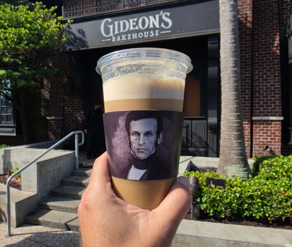 Hand holding a cup of iced coffee with Lincoln portrait sleeve outside Gideon's Bakehouse storefront
