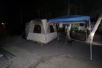 Night camping setup: beige tent under a blue canopy with string lights, folding chairs, and a trailer in the background.