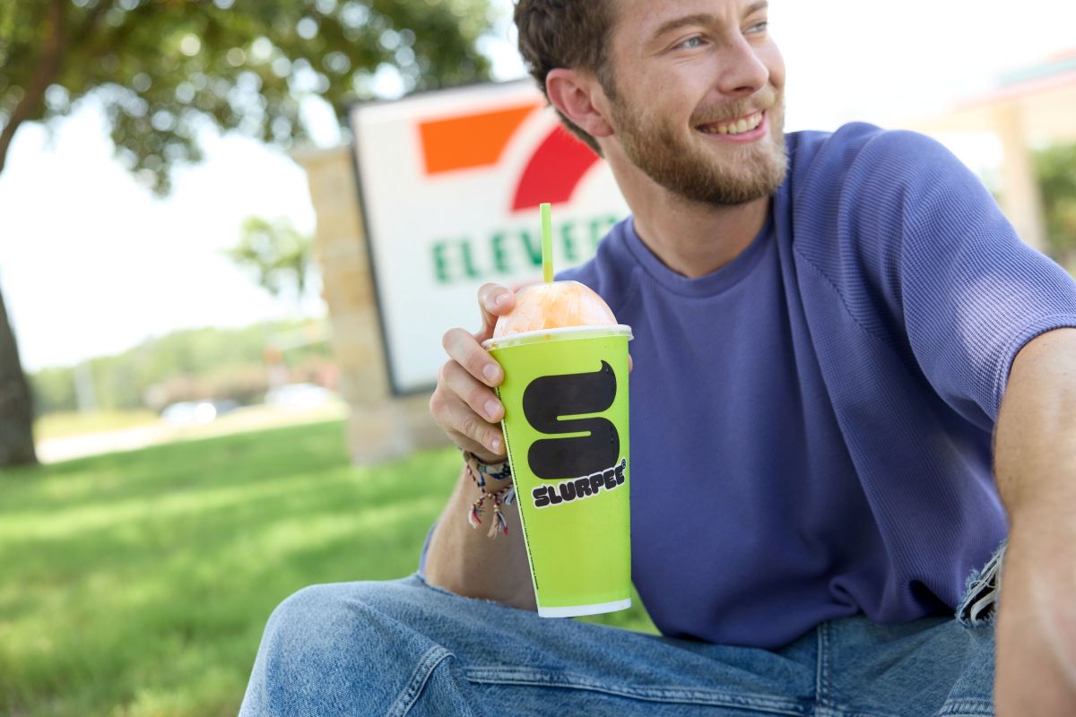 Smiling man sitting on grass outdoors, holding a bright green Slurpee cup with a straw for Slurpee Drink Happy Hour promotion