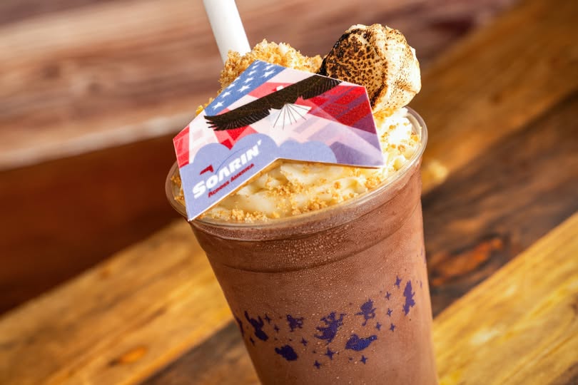 S'morin’ Across America Milk Shake in a tall glass topped with whipped cream, a cookie, and a decorative flag toothpick on a wooden table.