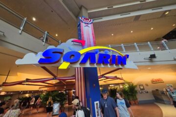Entrance to a theme-park ride with a large blue 'Soarin'' sign over fluffy white clouds and a patriotic column, with visitors walking underneath.