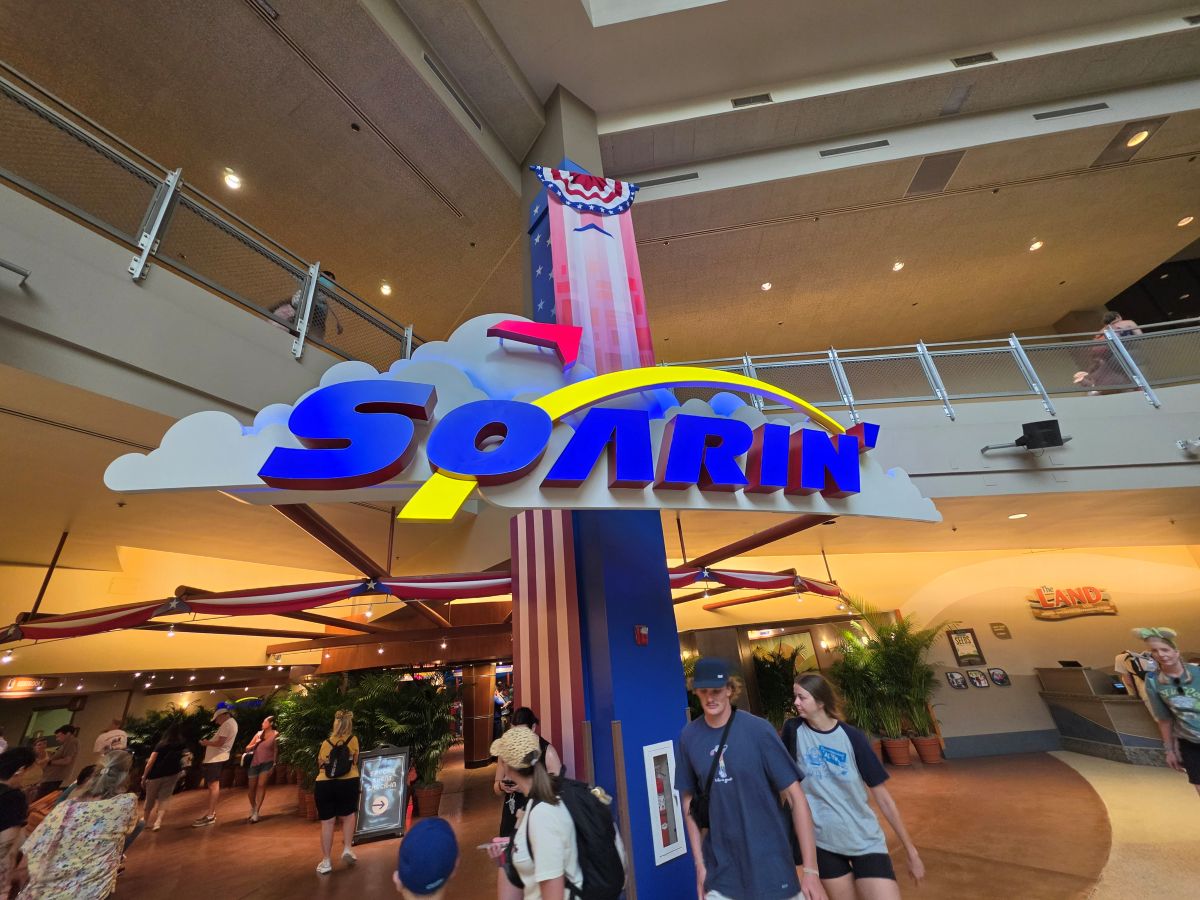 Entrance to a theme-park ride with a large blue 'Soarin'' sign over fluffy white clouds and a patriotic column, with visitors walking underneath.