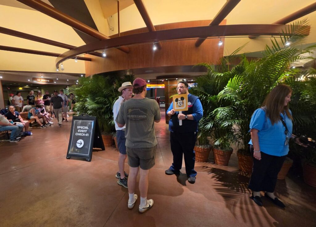 Guests in a lobby lined up for event check-in; a staff member holds a Mickey Mouse sign near tropical plants and a 'Special Event Check-In' sign with an arrow.