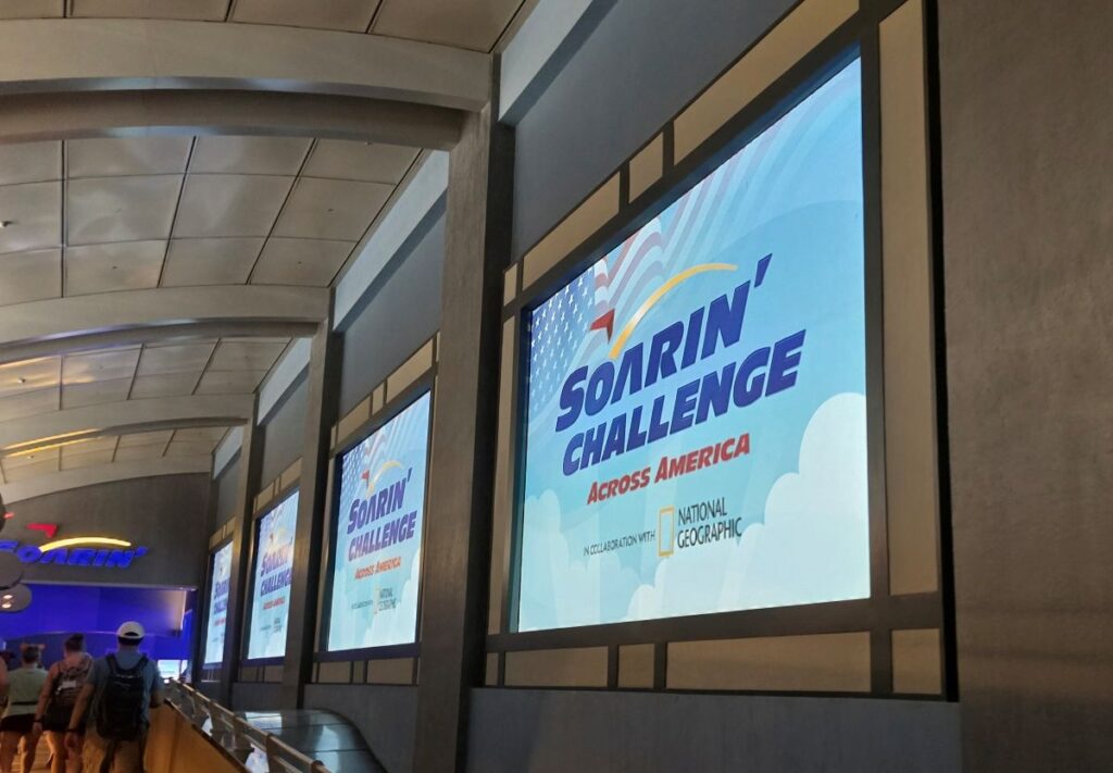 Indoor walkway with large blue posters reading 'Soarin' Challenge Across America' and a National Geographic logo along a glass-fronted wall, with people walking nearby.