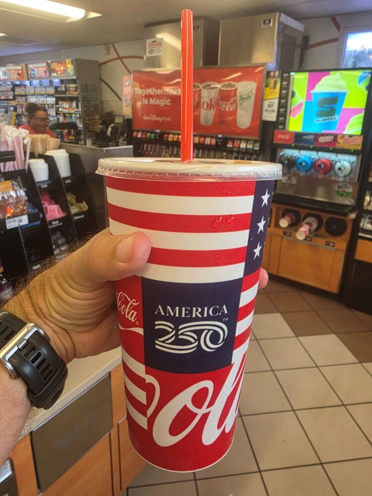 Person holding a patriotic Coca‑Cola cup with American flag design and 'America 250' in a convenience store.