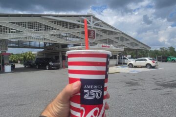 Person holding a red, white, and blue Coca‑Cola cup with a straw in front of a Speedway gas station for Speedway drink deals promotion