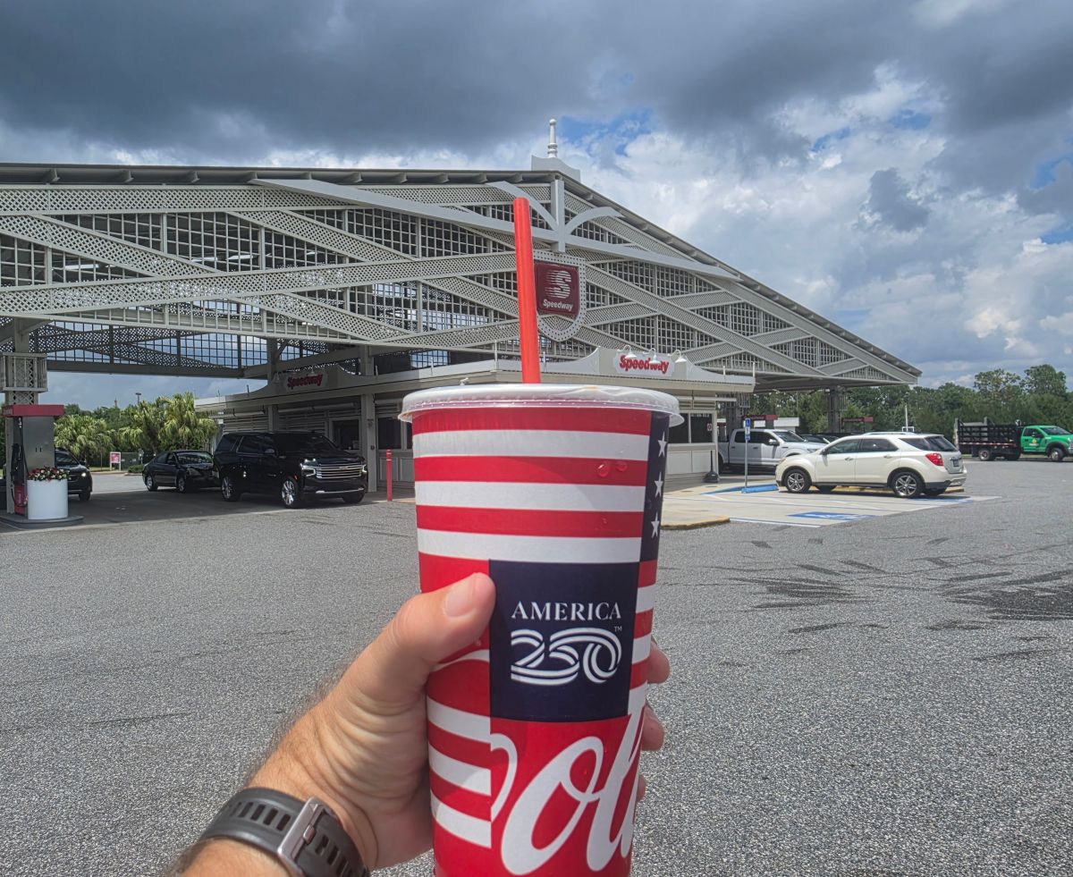 Person holding a red, white, and blue Coca‑Cola cup with a straw in front of a Speedway gas station for Speedway drink deals promotion