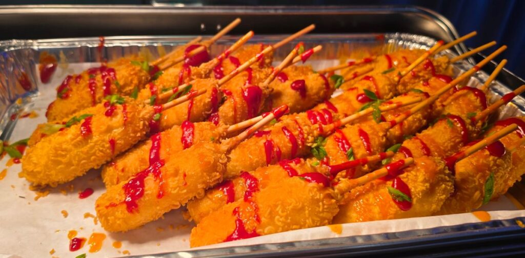 Tray of crispy corn dog skewers drizzled with red sauce and garnished with herbs on a serving tray.