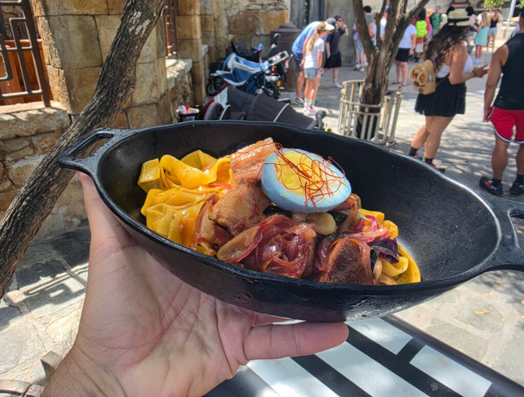 Hand holds a Puffer Pig Pasta black skillet filled with yellow pasta and seafood in the foreground, with a busy stone-paved street and tourists in the background.