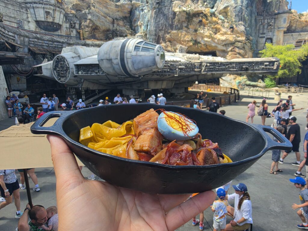Hand holding a black bowl of chips and meat in the foreground with a large sci‑fi spaceship ride and rocky backdrop in a crowded theme park.nNo. 1 Alt text for accessibility and context.