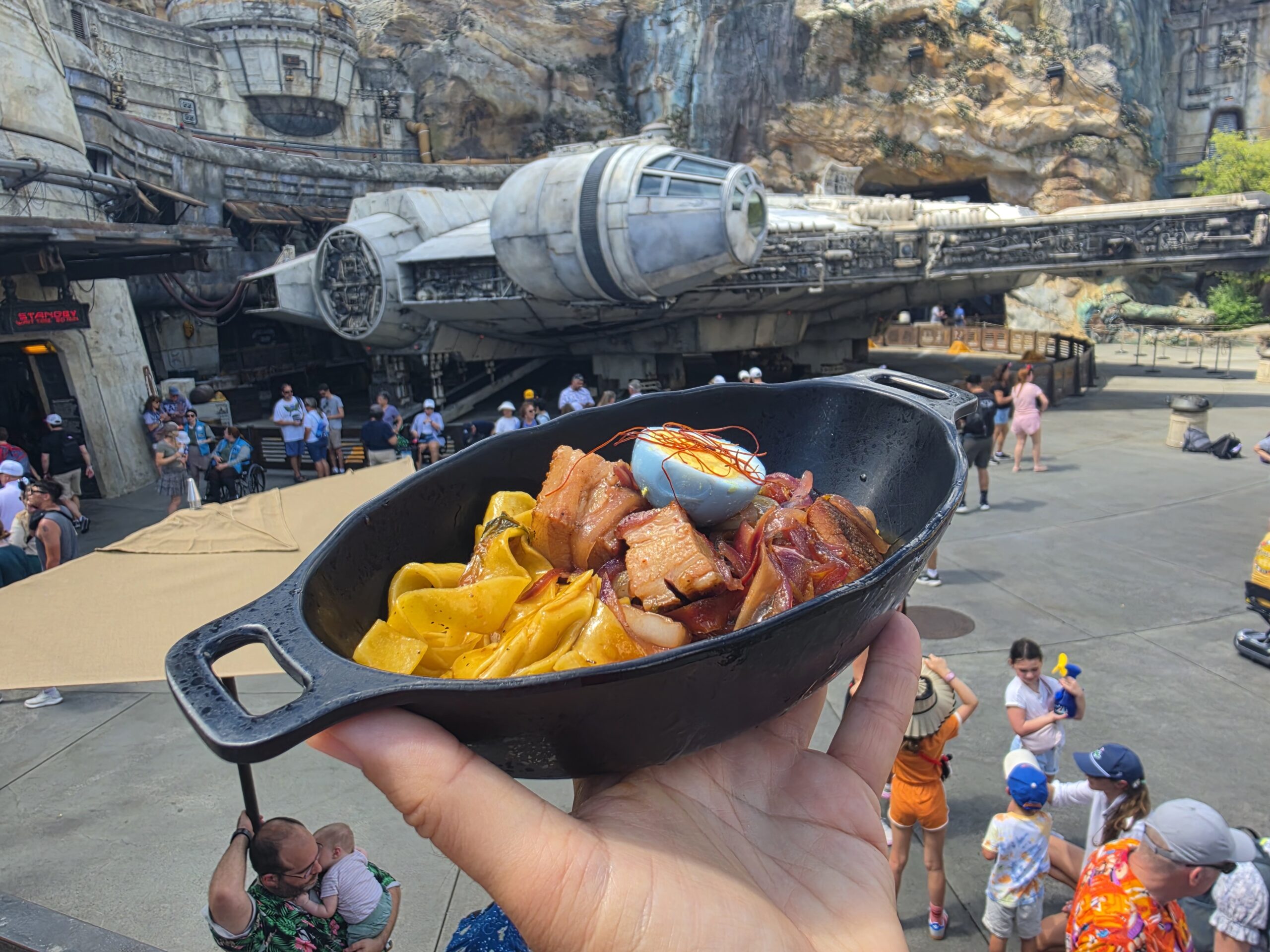 Close-up of a bowl of colorful pasta (yellow and reddish pieces) being held up, with a themed park and the Millennium Falcon ride in the background.