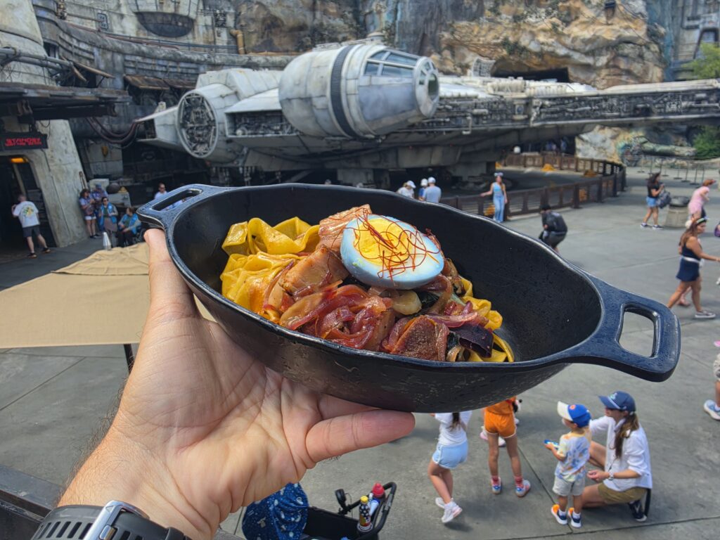 Hand holding a Puffer Pig Pasta skillet of colorful pasta with peppers, with the Millennium Falcon ride in the background at a theme park.
