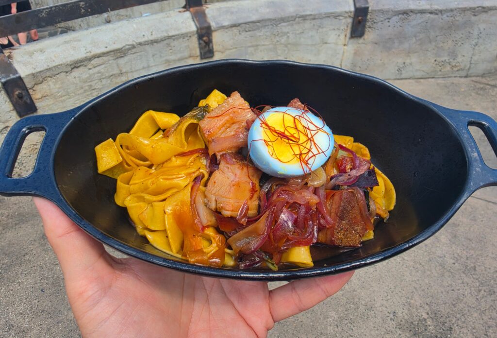 Hand holds a Puffer Pig Pasta black skillet filled with yellow pasta, sliced cured meat, and a blue egg topped with red threads outdoors.