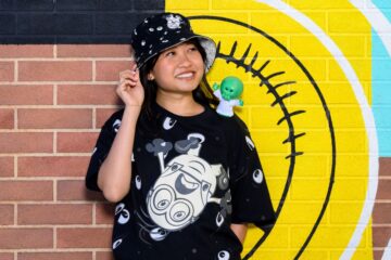 Smiling woman in a black graphic T-shirt and bucket hat stands against a yellow mural with a green alien toy perched on her shoulder.