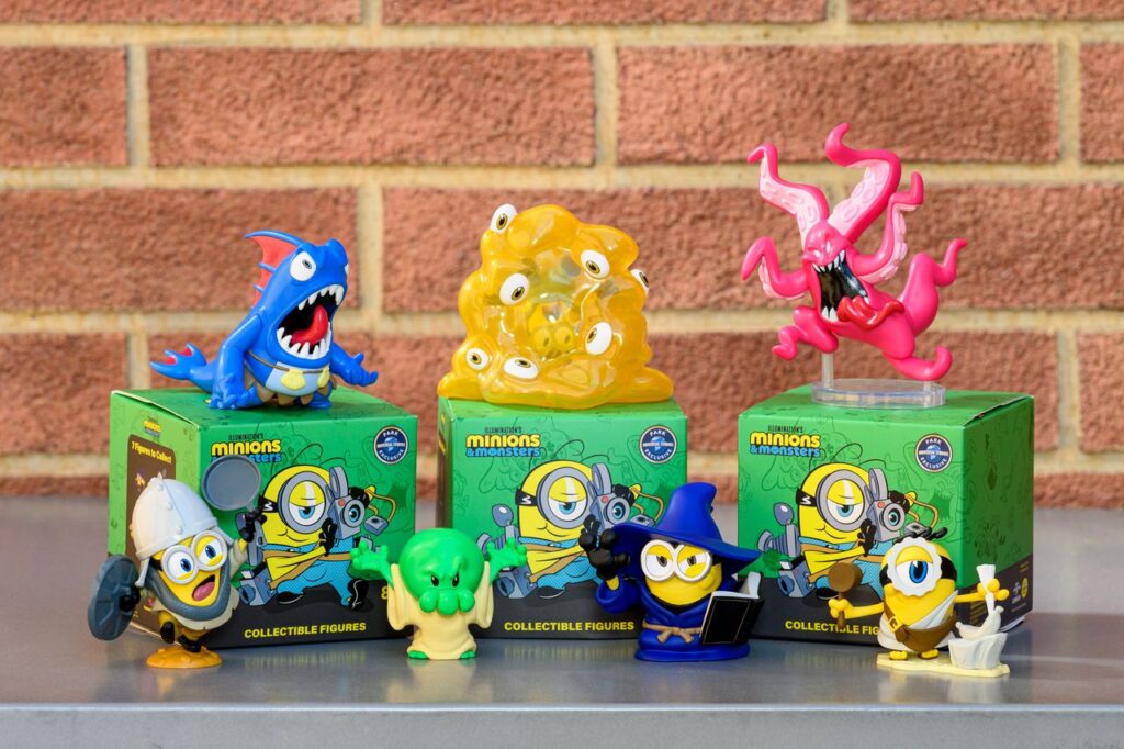 Group of Minions collectible figures arranged on green Minions boxes in front of a brick wall, with a blue shark-like monster, a yellow gelatinous blob, and a pink tentacled creature on top.