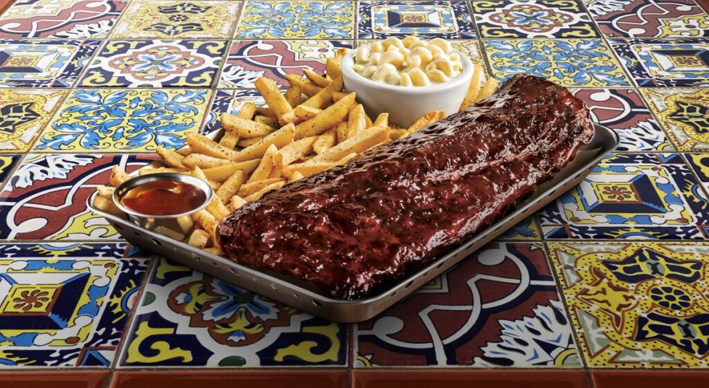 Barbecue ribs on a metal tray with seasoned French fries, macaroni salad, and barbecue sauce on a colorful tiled table.