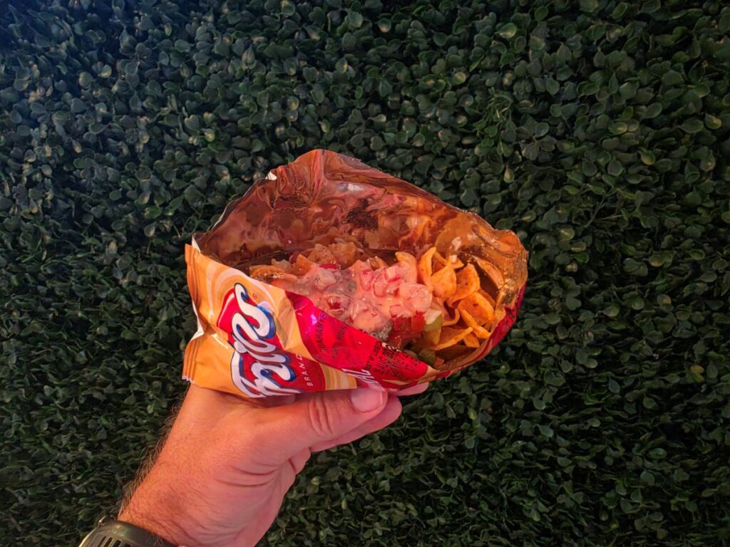 Hand holding an open bag of Fritos corn chips, chips spilling out, against a leafy hedge background and outdoors.