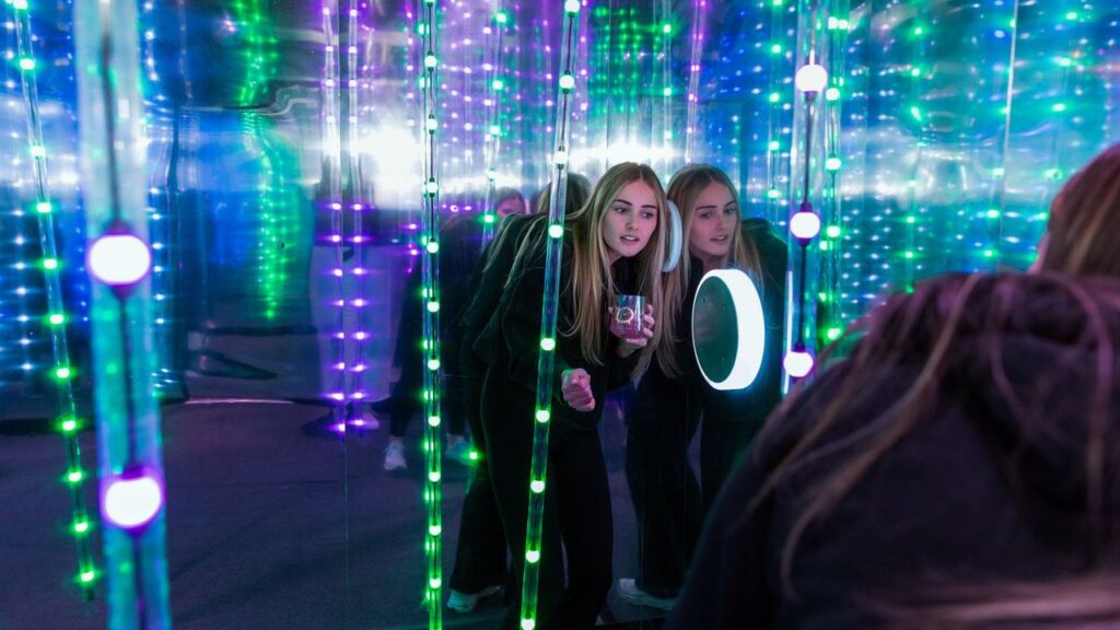 Two young women in a mirrored, neon-lit room with green and purple LED strands, looking at a glowing circular light panel.