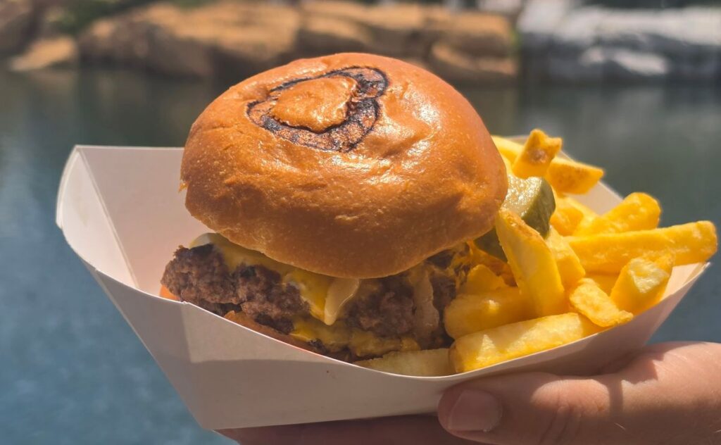 Richter's Burger Co Review photos showing a burger with cheese in a soft bun, served in a paper boat with a side of French fries, held by a hand near a lake.