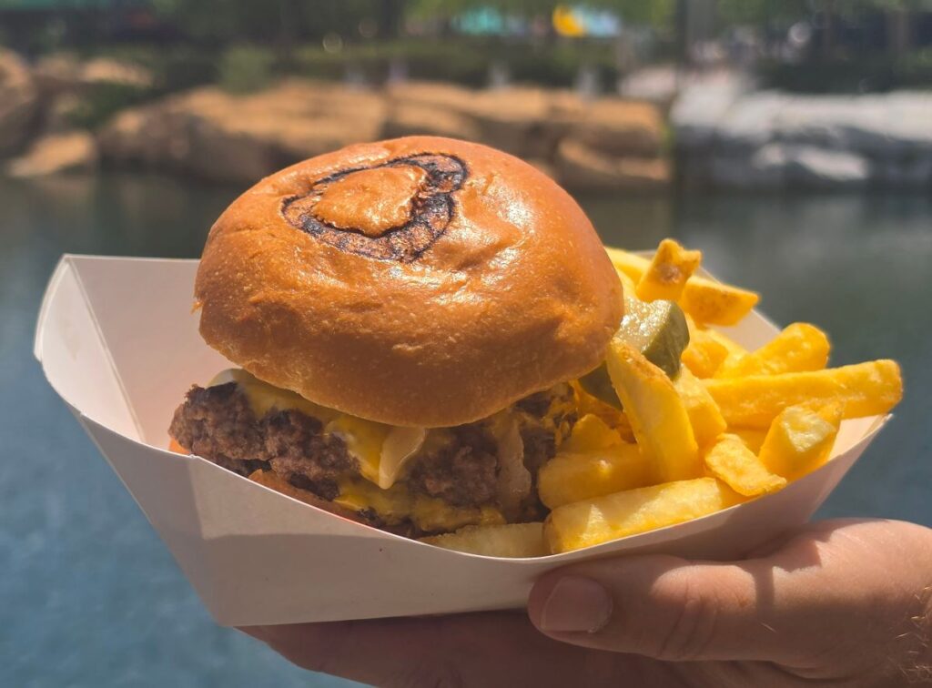 Burger with cheese and fried onions in a bun, held in a white tray with golden fries, outdoor waterfront scene in the background for a Richter's Burger Co Review