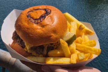 Burger with cheese and pickles in a bun, served with thick-cut fries on a white tray over blue water background for Richter's Burger Co Review