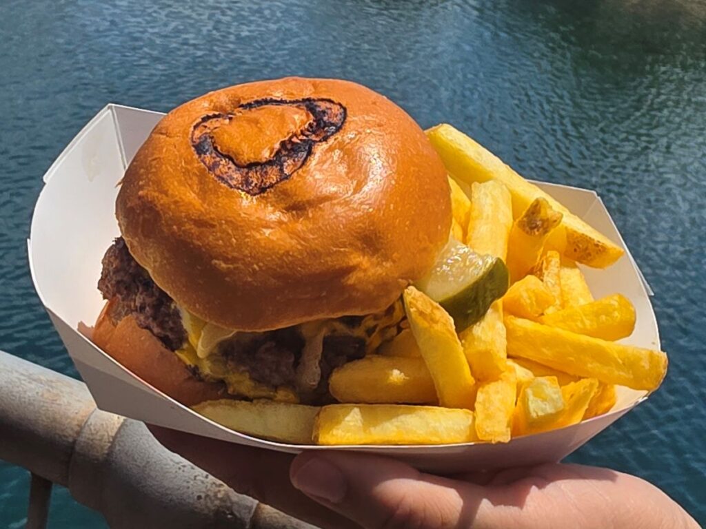 Burger with melted cheese in a glossy brioche bun and a side of French fries on a white tray by the water edge for Richter's Burger Co Review.