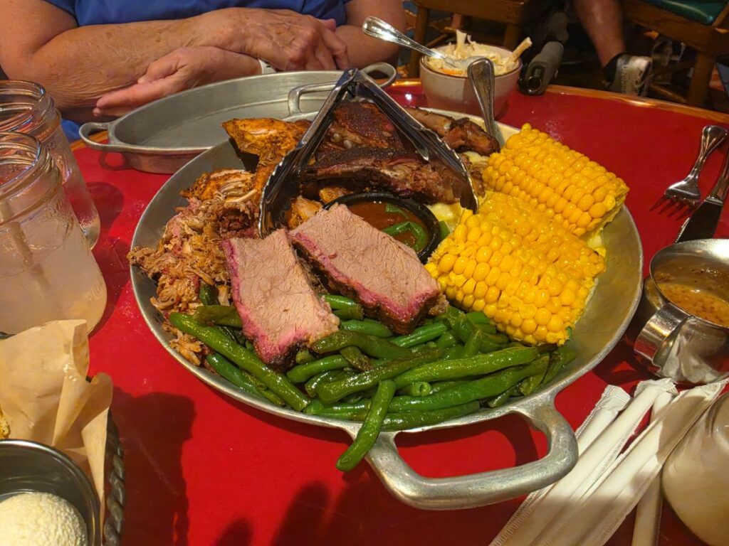 Smoked barbecue platter with sliced brisket, pulled pork, green beans, and corn on the cob on a metal serving tray for Whispering Canyon Cafe dinner