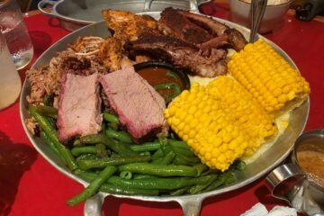 Metal serving platter with sliced brisket, pulled pork, green beans, two pieces of corn on the cob, and a side of barbecue sauce on the table for Whispering Canyon Cafe dinner