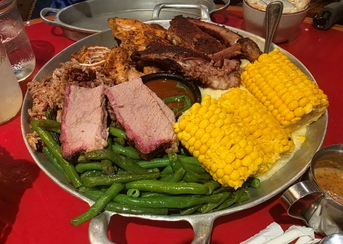 Metal serving platter with sliced brisket, pulled pork, green beans, two pieces of corn on the cob, and a side of barbecue sauce on the table for Whispering Canyon Cafe dinner